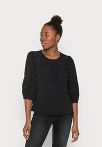 Blusa preta com mangas bufantes, detalhes em renda e um padrão texturizado. Possui um decote redondo e um corte descontraído, combinada com jeans escuros.