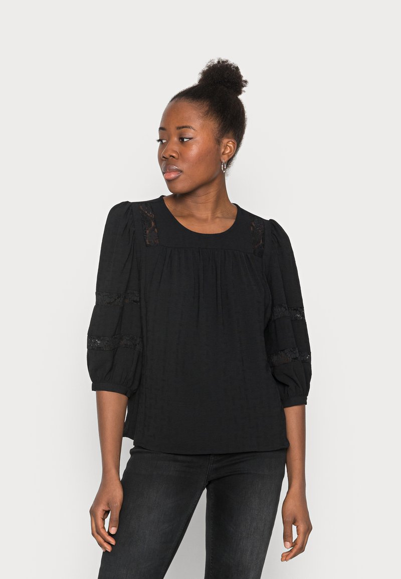 Blusa preta com mangas bufantes, detalhes em renda e um padrão texturizado. Possui um decote redondo e um corte descontraído, combinada com jeans escuros.