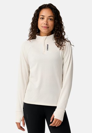 Mujer con cabello rizado que lleva una camiseta blanca de manga larga con cremallera hasta un cuarto y leggings negros, de pie sobre un fondo liso.