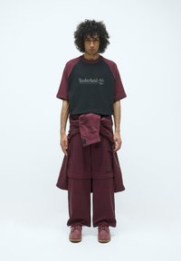 Herrenoutfit mit einem schwarz-burgunderfarbenen kurzärmeligen Hemd und passenden lockeren Hosen, hergestellt aus weichem Stoff mit sichtbarem Logo-Detail.