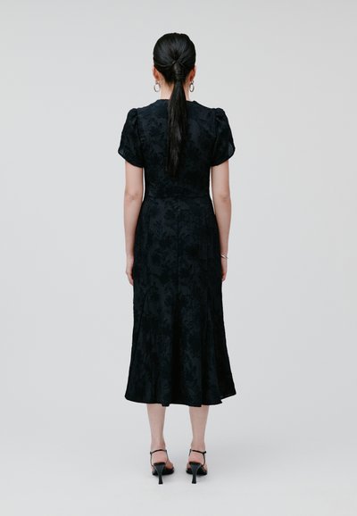 Schwarzes Kleid mit Blumendruck, kurzen Puffärmeln, mittel-langer A-Linien-Rock und strukturiertem Stoff. Das Model trägt schwarze hochhackige Sandalen.