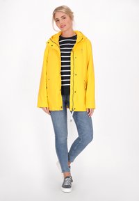 Gul vattentät jacka med huva, dragkedja fram och tryckknappar. Kombinerad med en marinrandig skjorta och blå jeans.