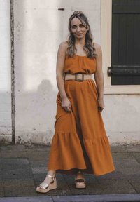 Rood-oranje, mouwloze maxi-jurk met een gelaagde rok en een gestructureerde stof, op de taille gepoft met een lichtgekleurde riem. Draag het met beige platform sandalen.