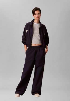 Junge Person steht mit den Händen in den Taschen, trägt schwarze Trainingsjacke und Hose mit weißen Streifen, graues kurz geschnittenes T-Shirt und beige Sneakers.