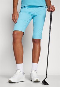 Ανοιχτό μπλε αθλητικά σορτς με λεία υφή, με σφιχτό σχέδιο. Συνδυασμένα με λευκά αθλητικά παπούτσια και κρατώντας ένα ρόπαλο γκολφ.