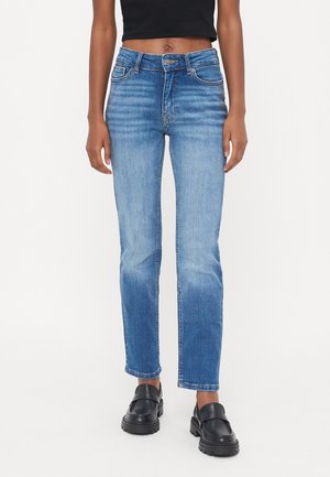 Persoon die mid-rise, recht gesneden blauwe spijkerbroek draagt, een zwarte cropped top en zwarte chunky loafers, staand tegen een effen witte achtergrond.