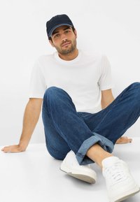 T-shirt bianca a maniche corte, jeans blu scuro, sneakers bianche e un cappellino blu navy con accenti azzurri. Pose casual seduta su uno sfondo bianco.