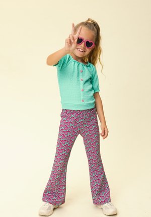 Meisje met hartvormige zonnebril, turquoise top met roze knopen, patroonbroek en witte sneakers, maakt met één hand een peace-teken.