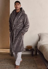 Grå plysjakke med ribbet tekstur, bred krave og bælte, båret over stribede pyjamabukser. Blødt stof og afslappet design.
