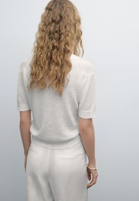 Lyhythihainen, vaaleanharmaa neulepaita, jossa on ribbireunat helmassa ja hihoissa, käytetty yhteensopivien vaaleiden housujen kanssa, nähtynä takaa.