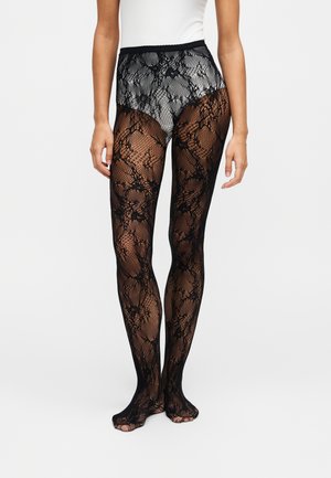 Femme portant des collants fins transparents à motif floral noir en dentelle et un haut blanc, debout les pieds rapprochés devant un fond uni.