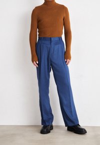 Maglione a collo alto marrone a coste e pantaloni larghi blu dalla texture liscia. Le calzature sono nere con una suola spessa. Stile minimalista.