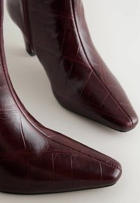 Burgundy nahka nilkkurit kiiltävällä pinnalla, kärkien muoto on terävä ja pinnassa on teksturoitu krokotiilikuosi. Kengissä on matala korko ja sivuvetoketju.