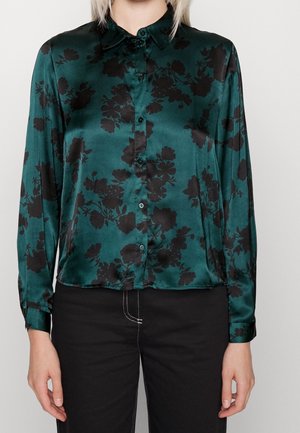 Blouse en satin bleu sarcelle avec motif floral noir, devant boutonné, manches longues, portée avec un pantalon noir avec des détails de couture blanche.