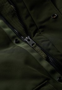 Tissu de veste vert foncé avec une fermeture éclair noire et des détails texturés verts, dotée d'un bouton-pression et d'accents tissés le long de la ligne de la fermeture éclair.