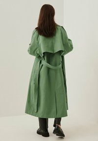 Długi zielony płaszcz trenchowy wykonany z lekkiego materiału, z paskiem w talii, dużym kołnierzem i warstwowym tyłem. Widoczne czarne botki poniżej.