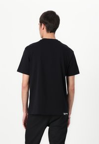 Camiseta de algodón negra de cuello redondo y mangas cortas. Textura suave y una etiqueta en la parte inferior izquierda. La vista trasera muestra un ajuste relajado.