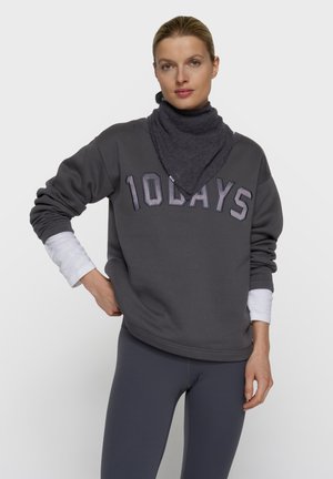Frau trägt dunkelgrauen Sweatshirt mit dem Text "10DAYS", grauen Hals Schal, weißes langärmeliges Shirt darunter und dunkelgraue Leggings, steht vor weißem Hintergrund.