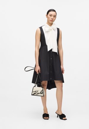 Kobieta w czarnej sukience bez rękawów z dużą białą kokardą, trzymająca małą białą torebkę i czarne sandały na obcasie.