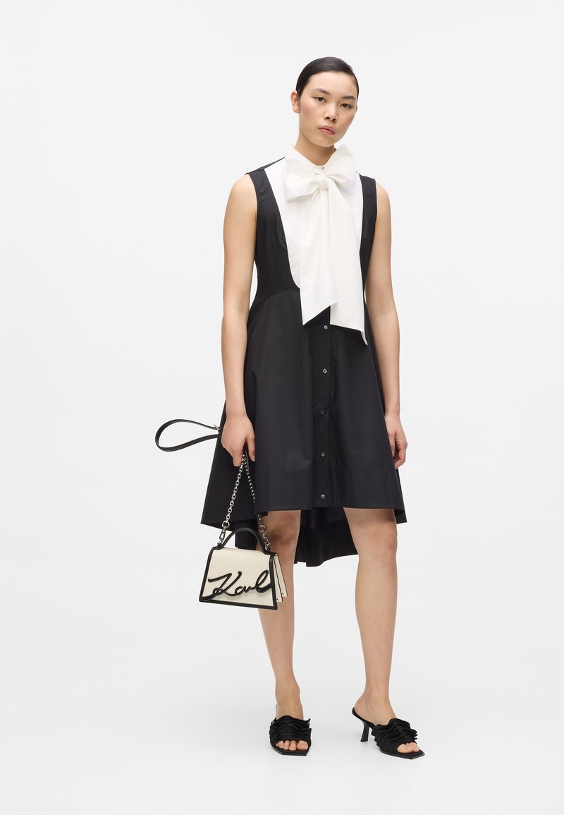 Γυναίκα που φοράει μαύρο αμάνικο φόρεμα με λευκό μεγάλο φουλάρι φιόγκο, κρατώντας μικρή λευκή τσάντα και μαύρα σανδάλια με τακούνι.