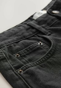 Jeans de mezclilla negros con un bolsillo frontal adornado con tachuelas de cobre, una textura sutil y costuras distintivas a lo largo de los bordes.