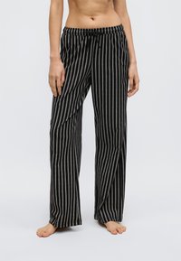 Person med blottad midja som bär svarta och vita löst sittande byxor med vertikala ränder, elastisk midja och dragsko, stående barfota på enfärgad bakgrund.