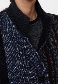 Abrigo de mezcla de lana que presenta un panel texturizado azul marino con un patrón de espiga, cuello grueso y detalle de botones negros. Capa interior gris visible.