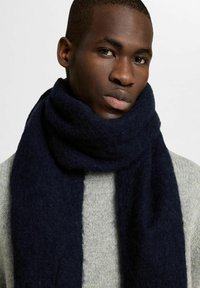Sciarpa di lana blu scuro drappeggiata attorno al collo, con una finitura morbida e testurizzata. Indossata con un maglione grigio chiaro a coste.