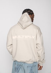 Homme aux cheveux tressés portant un sweat à capuche beige avec le texte « MULTIPLY APPAREL CLUB » au dos et un jean bleu clair avec des motifs cousus sur les poches.