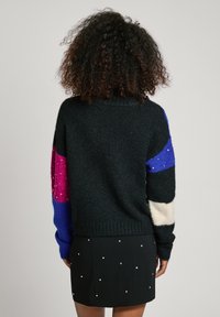 Schwarzer Pullover mit bunten gestreiften Ärmeln in Pink, Blau und Creme, mit weicher Textur und lockerer Passform, kombiniert mit einem schwarzen Rock.