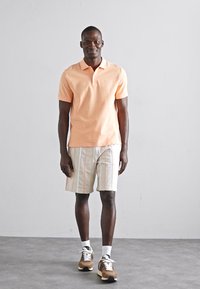 Lyst orange polo shirt, hvide shorts med lodrette striber og brune sneakers med hvide snørebånd. Mand står med neutral baggrund.