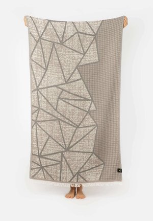 Béžová a sivá geometricky vzorovaná deka so strapcami držená osobou, nohy viditeľné pod ňou na bielom pozadí.