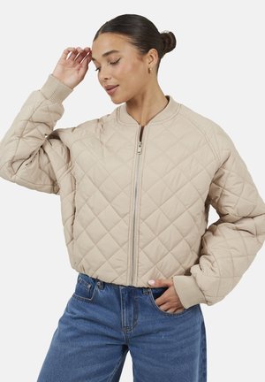 Donna con capelli scuri raccolti in uno chignon indossa una giacca trapuntata beige con cerniera e jeans blu, posa con una mano sul fianco e l'altra vicino alla testa.