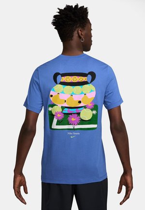 Man in een blauwe Nike tennis T-shirt met een kleurrafische afbeelding van een vaas, tennisballen, peren en roze bloemen op de achterkant.