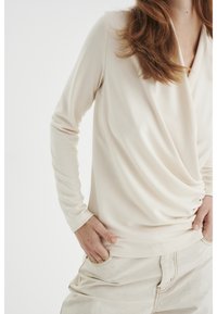 Kvinna med rödbrunt hår som bär en krämfärgad långärmad topp och ljusa byxor med händerna i fickorna.