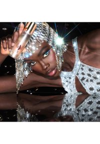 Glass serum bottle with metallic accents held against a shimmering silver outfit adorned with beads; mirrored surface reflects details.