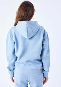 Felpa blu chiaro realizzata in morbido tessuto, con un ampio cappuccio, polsini e orlo a coste. Tessuto liscio senza motivi visibili.