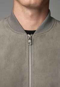 Gros plan sur le cou et la poitrine d'une personne portant une veste en daim grise avec une fermeture éclair argentée et un col côté par-dessus une chemise noire.