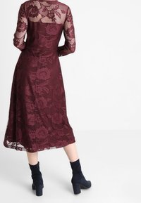 Burgundské krajkové šaty s dlouhými rukávy, květinovým vzorem a střední délkou. V kombinaci s tmavě modrými kotníkovými botami na podpatku.