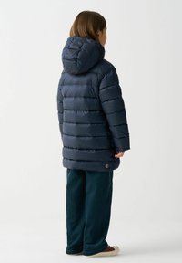 Námornícka modrá puffová bunda s horizontálnym prešívaním, integrovanou kapucňou a kruhovým logom na dolnom leme, skombinovaná s tmavozelenými nohavicami a béžovými topánkami.