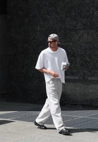 Bela oversize majica, svetlosive široke hlače, crne cipele, siva kapa, naočare za sunce, drži piće i telefon, hoda po textured površini.