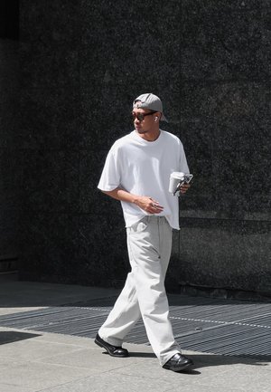 Witte oversized t-shirt, lichtgrijze wide-leg broek, zwarte schoenen, grijze pet, zonnebril, met een drankje en telefoon in de hand, wandelend over een textuur oppervlak.