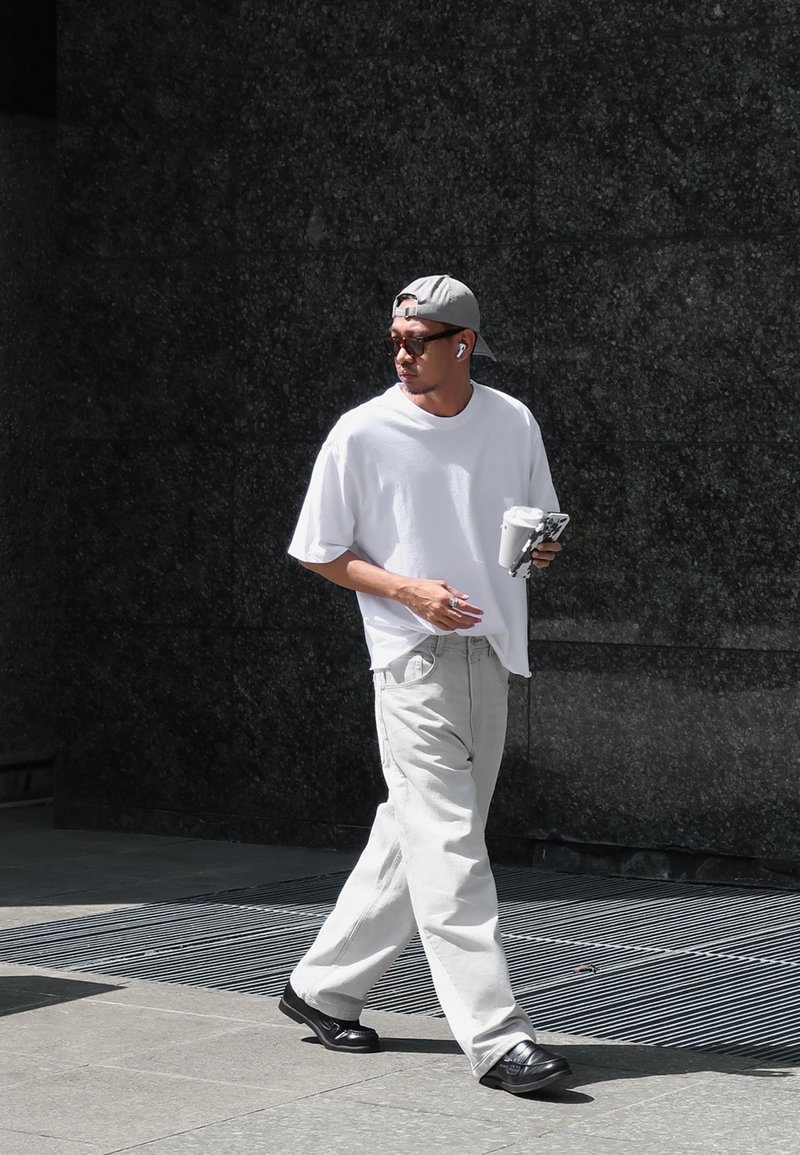 Bela oversize majica, svetlosive široke hlače, crne cipele, siva kapa, naočare za sunce, drži piće i telefon, hoda po textured površini.