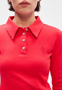 Polo roșu cu dungi, cu guler clasic, prezentând patru nasturi din piatră prețioasă transparentă și un mic logo brodat pe piept.
