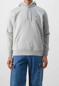 Felpa grigia in cotone con tasca a marsupio, cappuccio regolabile e piccolo logo sul petto. Abbinata a jeans cargo azzurri chiari.