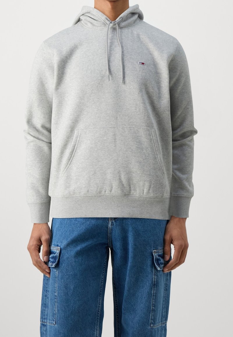 Felpa grigia in cotone con tasca a marsupio, cappuccio regolabile e piccolo logo sul petto. Abbinata a jeans cargo azzurri chiari.