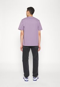 Moški obrnjeno stran, oblečen v enostavno sivoljubičasto majico, črne kavbojke in bele superge Adidas z modrimi črtami na beli podlagi.