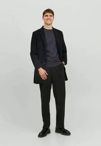 Sobretudo preto ajustado sobre um suéter escuro e calças pretas largas. O modelo está de pé de forma casual com as mãos nos bolsos, usando sapatos pretos.