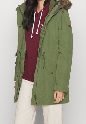 Femme portant un parka vert avec capuche en fourrure par-dessus un sweat à capuche bordeaux et un pantalon blanc, debout devant un fond uni.