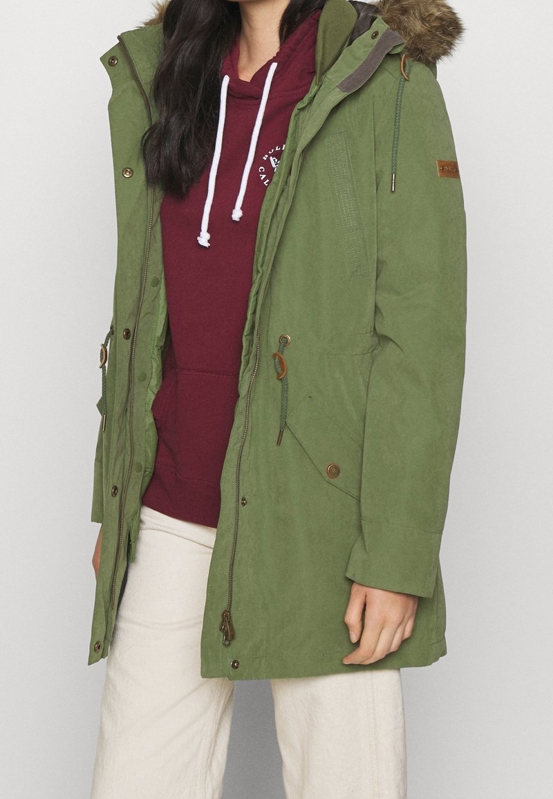 Femme portant un parka vert avec capuche en fourrure par-dessus un sweat à capuche bordeaux et un pantalon blanc, debout devant un fond uni.